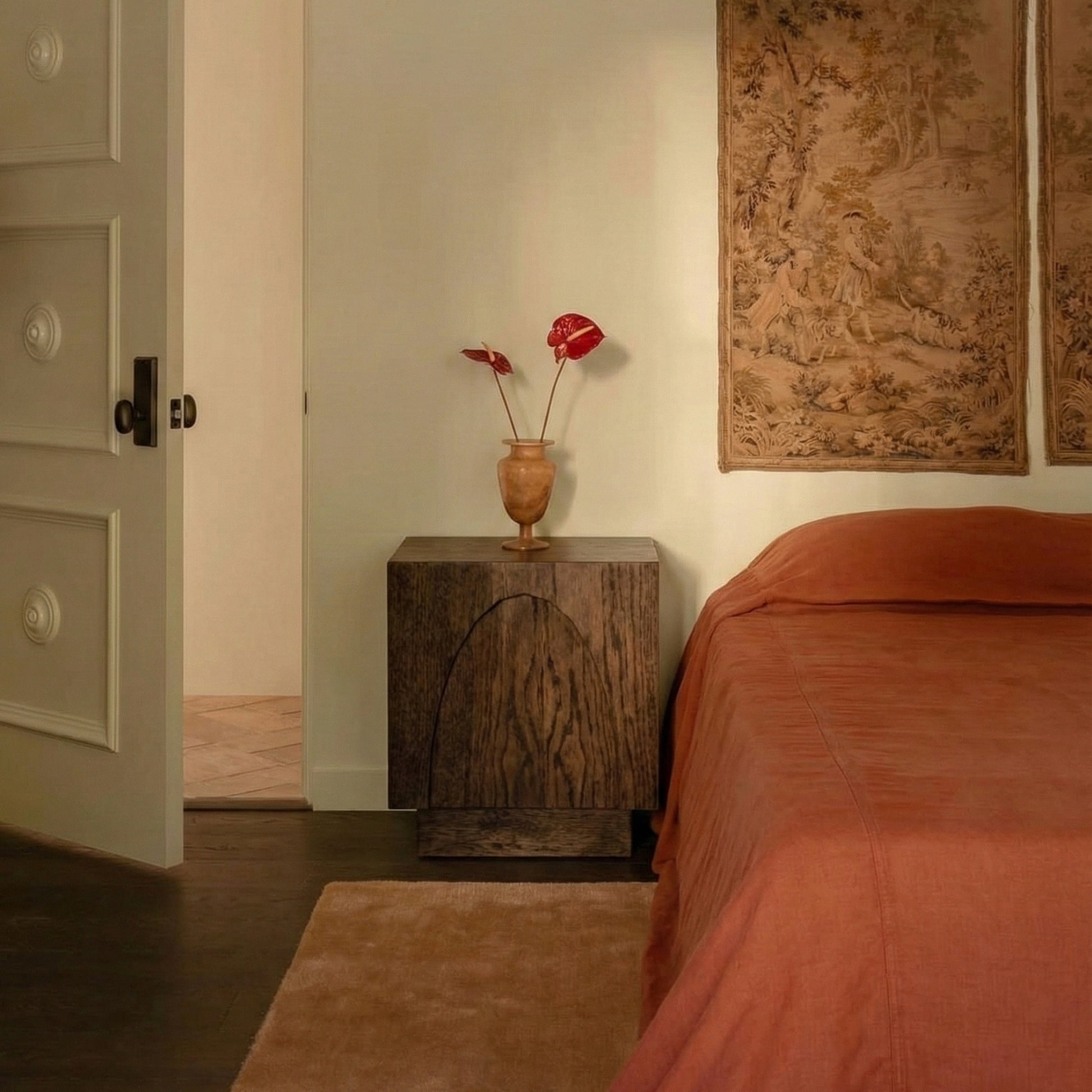 Bedroom with wooden nightstand, vase with red flowers, and large tapestry on wall.